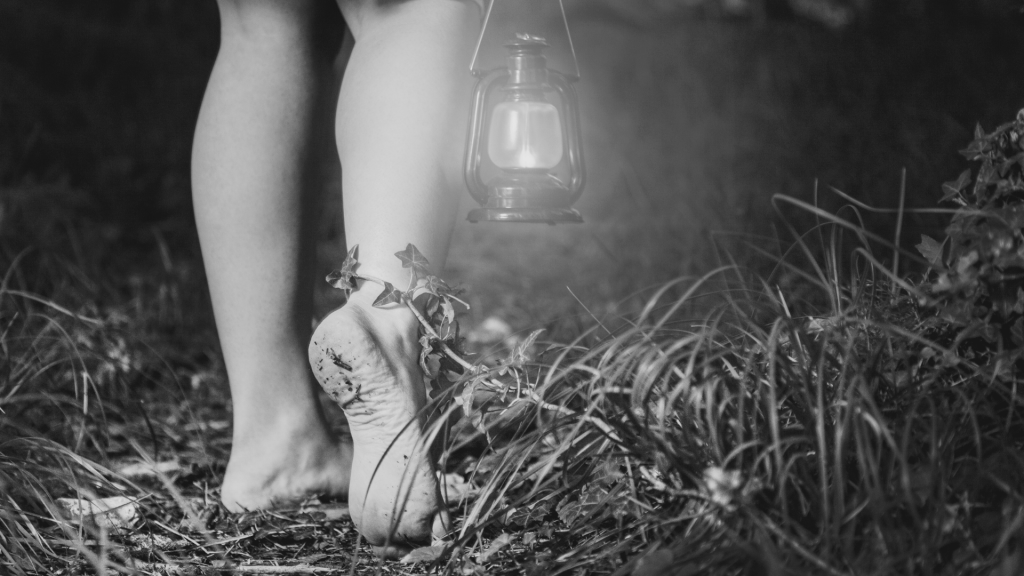 photo noir et blanc de pieds nus d'une femme marchant dans la nature. Elle tient une lampe ancienne car il fait nuit.