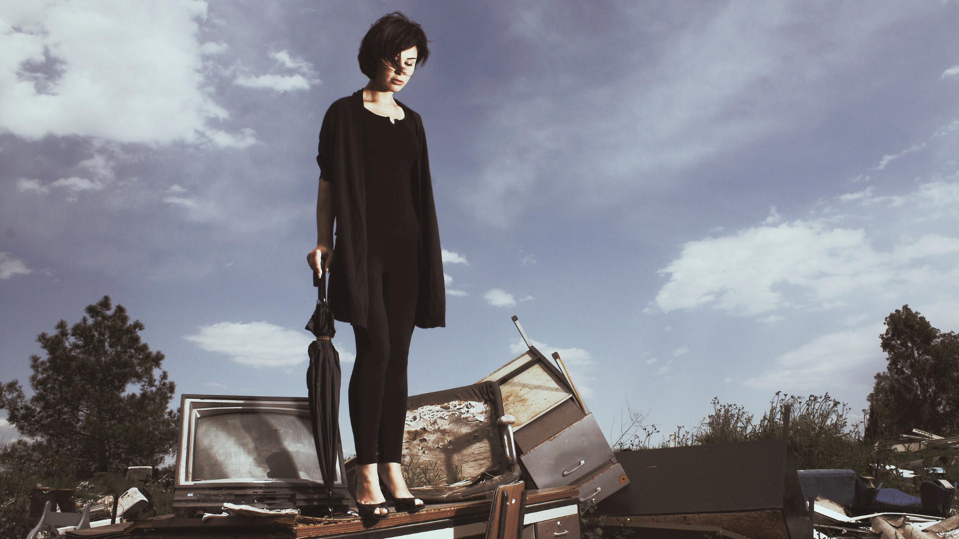 une femme vêtue de noir, en talon haut, est juchée sur un monticule de déchets (vieille télé cathodique, mobilier de bureau...) Elle a la tête inclinée vers le bas comme pour regarder où elle pourrait bien poser les pieds.