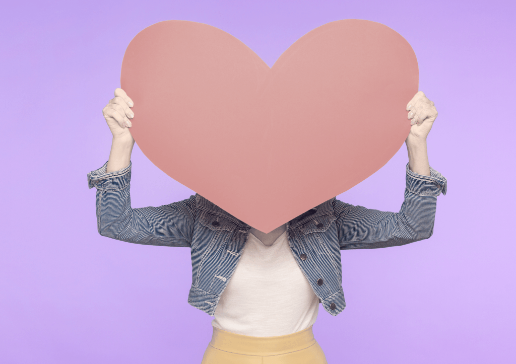 Une personne debout tient devant son visage un gros cœur rouge en papier cartonné qui masque son visage et ses épaules